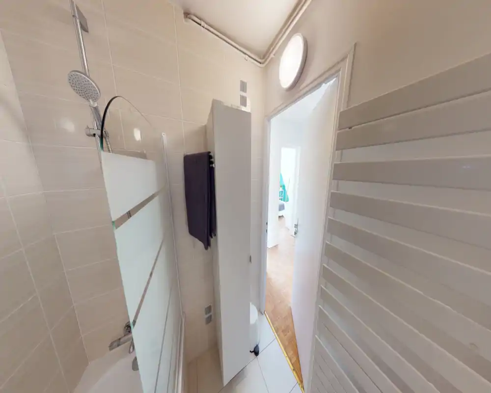 Compact modern bathroom with tiled walls, a bathtub with a glass shower screen and an overhead shower. A tall white storage cabinet and towel are visible near the doorway.