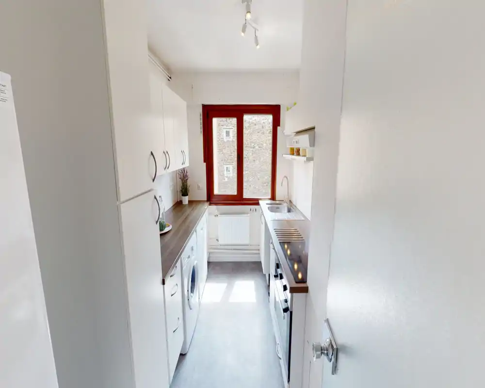 Bright compact kitchen with white cabinets, wood-effect countertops, built-in oven and sink under a large window that provides natural light.