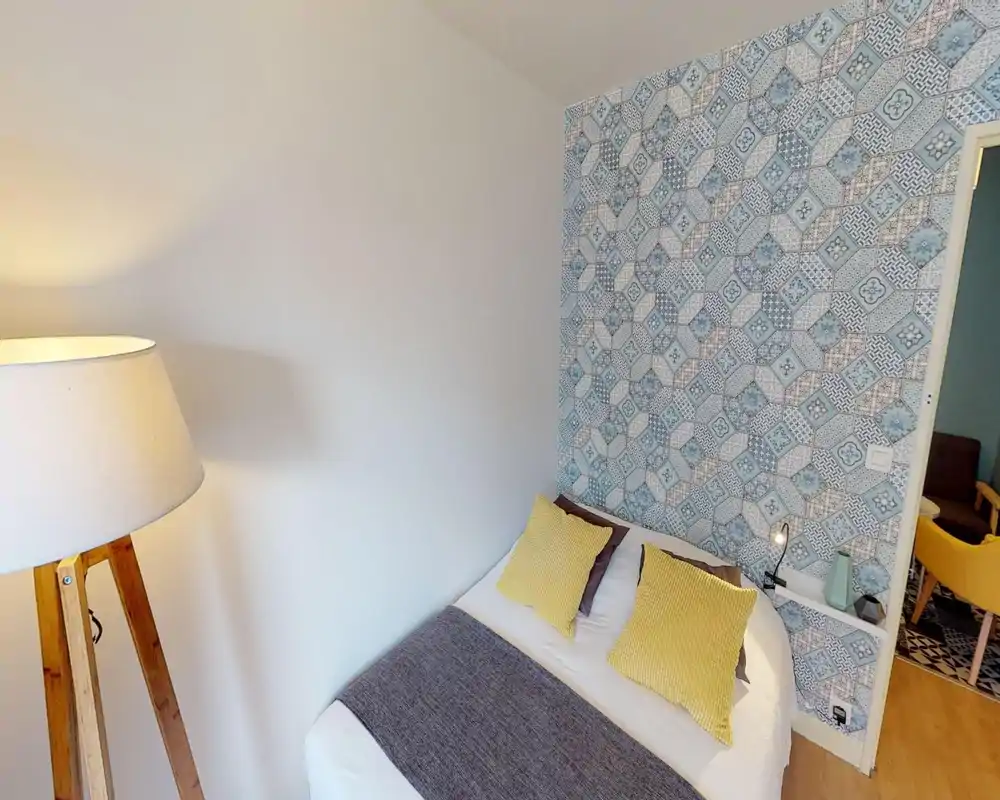 Camera da letto accogliente con un letto matrimoniale con lenzuola bianche, cuscini gialli a contrasto e una parete decorativa con piastrelle blu; lampada da terra calda e comodino curato completano l'ambiente.