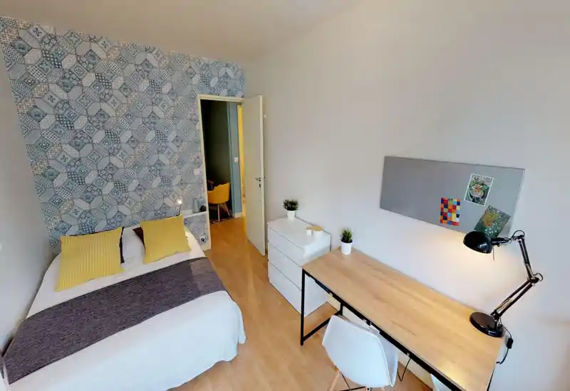 Camera da letto luminosa e ben allestita con parete decorativa a motivo, letto matrimoniale con cuscini gialli e una scrivania ordinata — perfetta per lavorare e riposare.