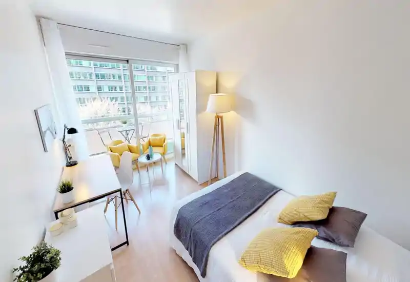 Camera da letto moderna e luminosa con letto matrimoniale, cuscini decorativi e plaid. Ampia finestra a tutta altezza con balcone e zona salotto, armadio, scrivania e una lampada da terra che crea un’atmosfera accogliente.