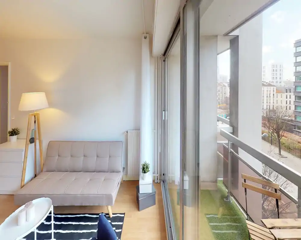 Bright modern living room with a compact sofa, floor lamp and sliding glass doors opening to a small balcony — ideal for city rentals seeking a cozy, well-lit lounge.
