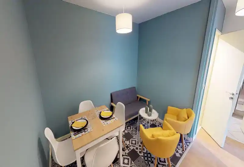 Cozy living/dining nook with a small wooden table, white chairs, a compact sofa and two yellow armchairs — stylish teal walls and patterned floor tiles give the space character.