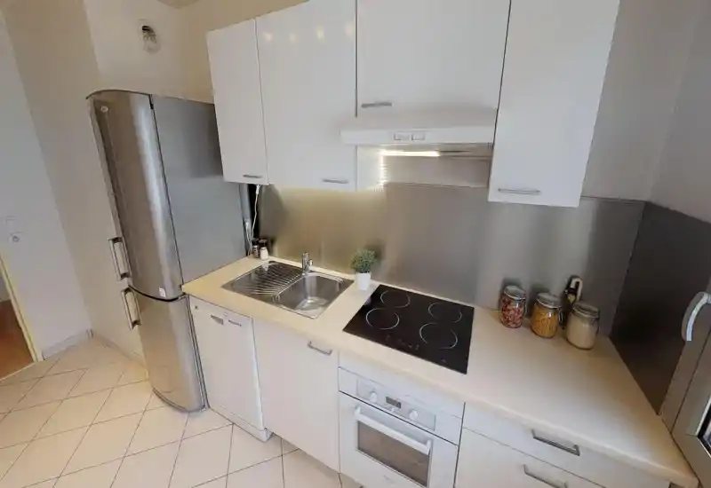 Modern compact kitchen with stainless steel refrigerator, built-in oven and ceramic cooktop, under-cabinet lighting and a clean countertop with jars and a small plant.
