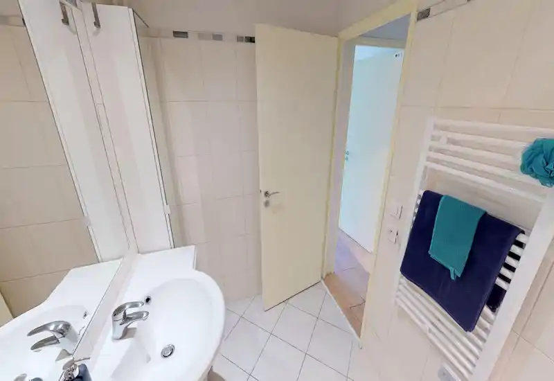 Clean, well-lit bathroom with a white sink, mirror and a heated towel rail holding folded towels. Neutral tiles and soft lighting create a fresh, functional space.