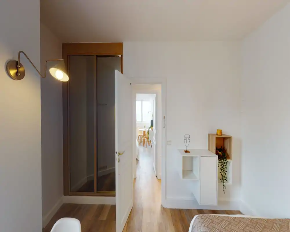 Angolo camera luminoso e minimal con pavimento in legno caldo, lampada da parete, mensole sospese e letto parzialmente visibile in primo piano. Stile pulito e luce naturale che rendono lo spazio accogliente.