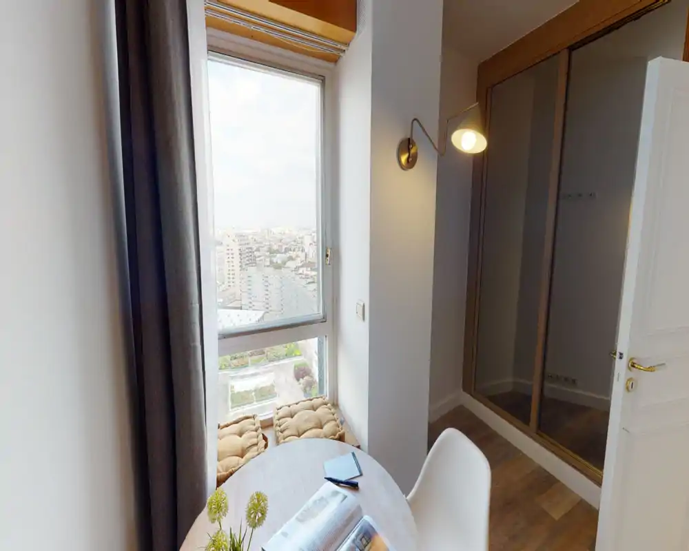 Angolo soggiorno luminoso con ampia finestra, tavolino rotondo, cuscini sul davanzale e una sedia comoda — perfetto per leggere o godersi la vista sulla città.