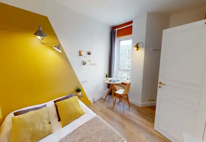 Camera da letto luminosa e alla moda con parete gialla a contrasto, letto comodo con cuscini testurizzati e piccolo tavolo vicino alla finestra — perfetta per un soggiorno accogliente o un angolo studio.