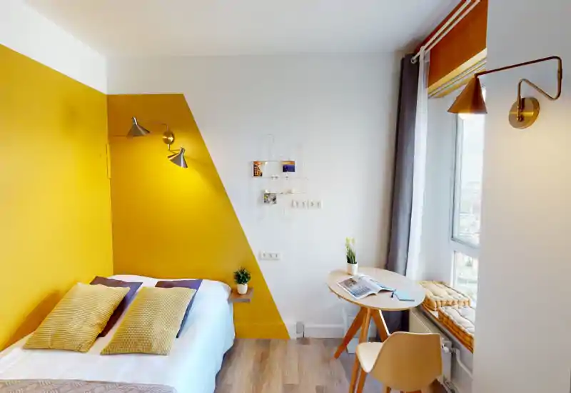 Camera da letto moderna e luminosa con parete accent giallo senape, letto matrimoniale con cuscini testurizzati, lampade da lettura e piccolo tavolo vicino alla finestra — accogliente e ben illuminata.