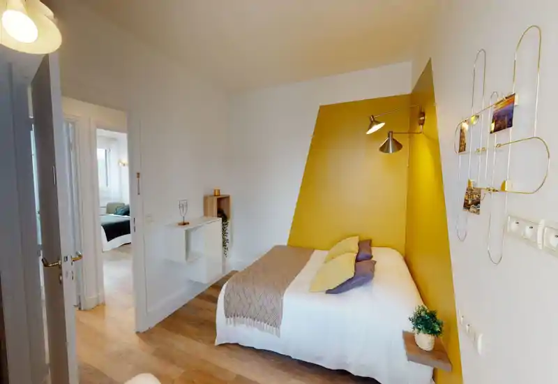 Camera da letto luminosa e di stile con parete accentata giallo senape, letto matrimoniale con biancheria bianca e cuscini sovrapposti, e pavimento in legno caldo — spazio notte moderno e ben allestito.