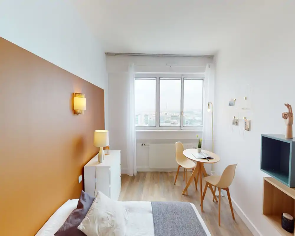 Chambre lumineuse et stylée avec un mur d'accent chaud, lit bien fait au premier plan et un petit coin table/bureau près de la fenêtre offrant une vue sur la ville.