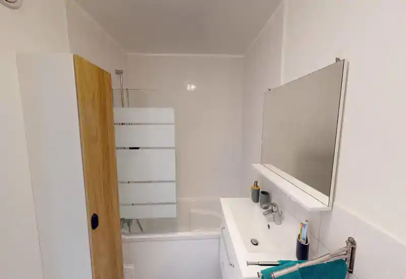 Clean, compact bathroom with a bathtub, glass shower screen, sink with vanity and a wall mirror. Neutral tiles and a tall storage cabinet make this bathroom practical and bright.