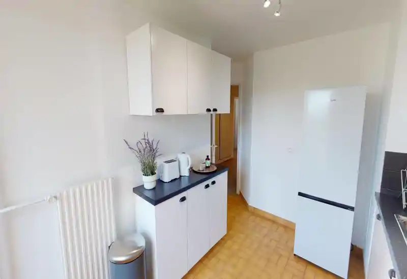 Bright, compact kitchen with white cabinets, a freestanding refrigerator and a tidy countertop featuring a kettle, toaster and potted plant — practical and well maintained.
