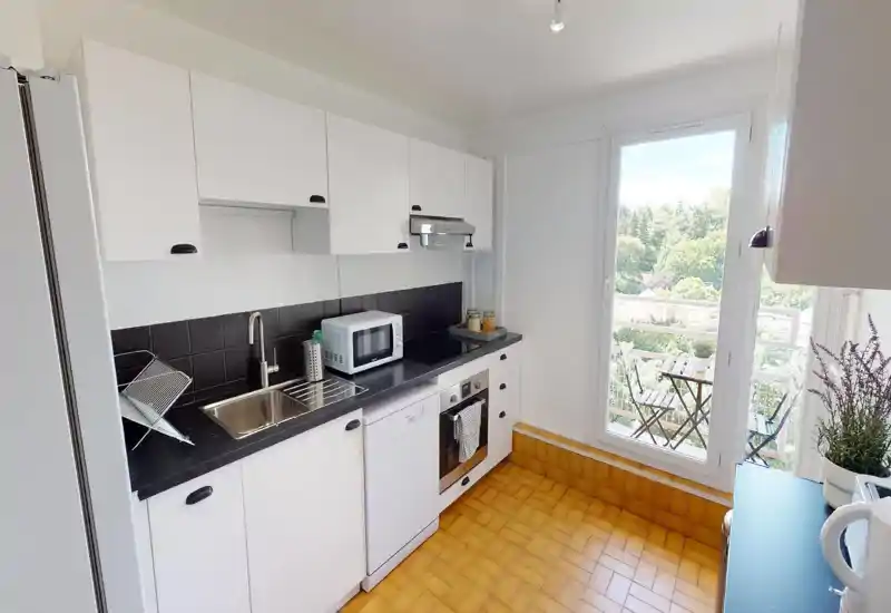 Bright modern kitchen with white cabinetry, a long countertop, skylight-style windows and a wooden breakfast table — airy and well lit.