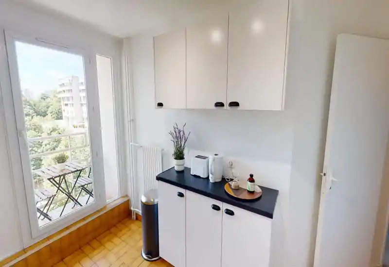 Compact, bright kitchenette with white cabinets, black countertop, and a small breakfast setup by a large glass door opening to a balcony.