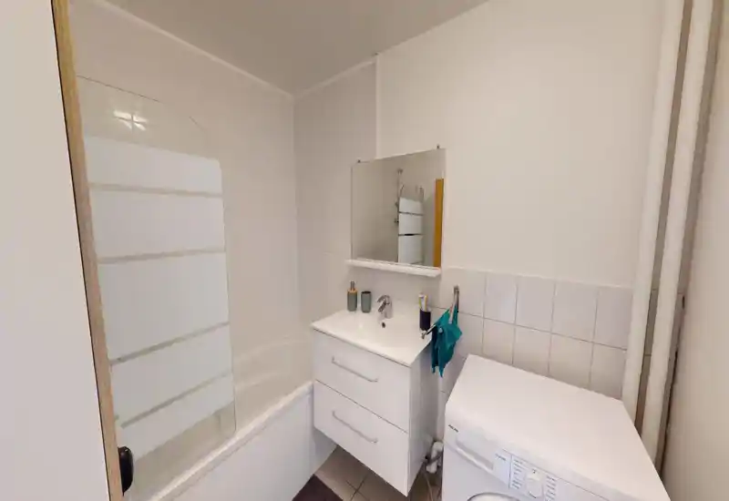Compact, well-lit bathroom with a bathtub/shower, vanity with sink and mirror, and a washing machine. Clean white tiles and modern fixtures create a fresh, practical space.