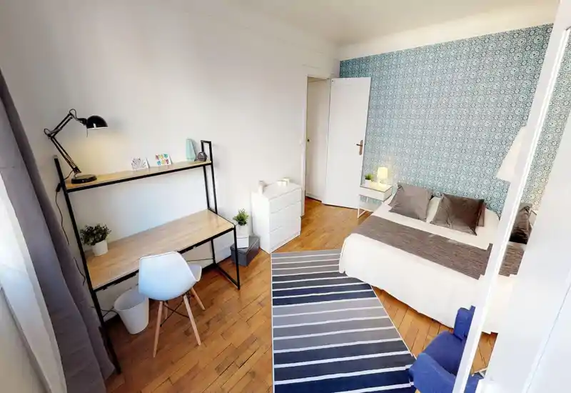 Camera da letto luminosa e ben arredata con letto matrimoniale davanti a una parete decorata, comodino con lampada, scrivania e sedia — perfetta per riposo e lavoro.