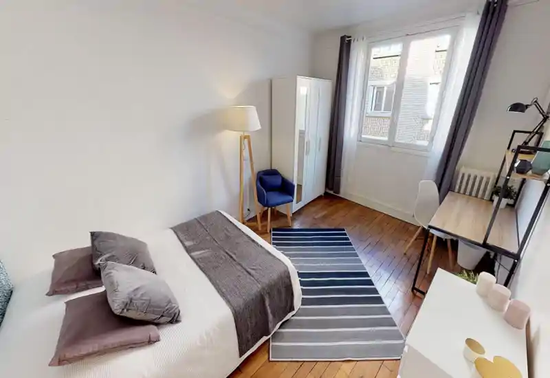 Camera da letto luminosa e ben arredata con letto matrimoniale, tappeto a righe, armadio e una piccola scrivania vicino alla finestra — ideale per una persona o una coppia. Pavimenti in legno, pareti neutre e tessili che creano un’atmosfera accogliente.