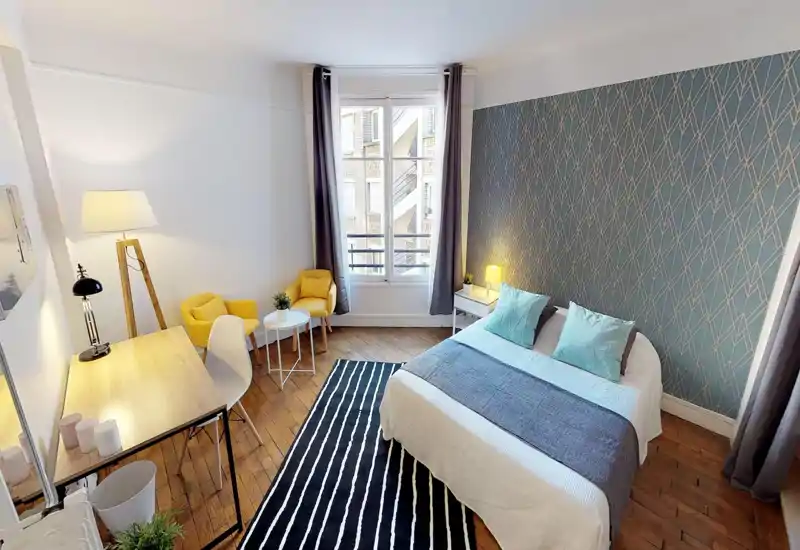 Camera da letto luminosa e di design con letto matrimoniale in biancheria bianca e cuscini turchesi, parete decorativa, pavimento in legno e angolo salotto vicino alla finestra.