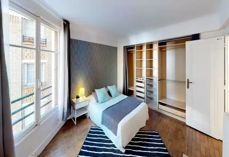 Camera da letto luminosa e stilosa con letto matrimoniale al centro contro una parete decorata, comodino con lampada, ampia finestra con luce naturale e guardaroba aperto a muro.