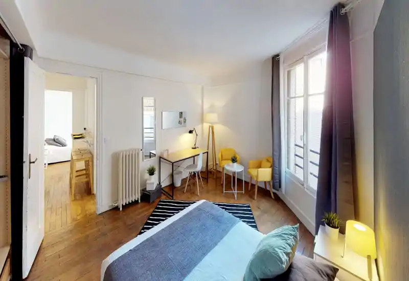 Camera da letto luminosa e curata, con ampia finestra, tessuti accoglienti e una scrivania. Il letto è ben visibile in primo piano, i pavimenti in legno e le luci calde valorizzano l'ambiente.
