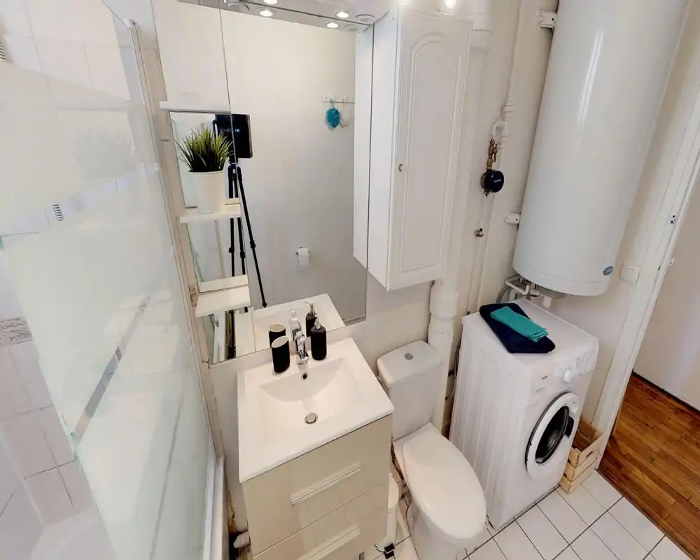 Bagno luminoso e ordinato con lavabo, WC, vasca con pannello in vetro satinato e una lavatrice sotto un grande scaldabagno. Piastrelle e mobili bianchi creano un ambiente funzionale e pulito, adatto ad affitti brevi.