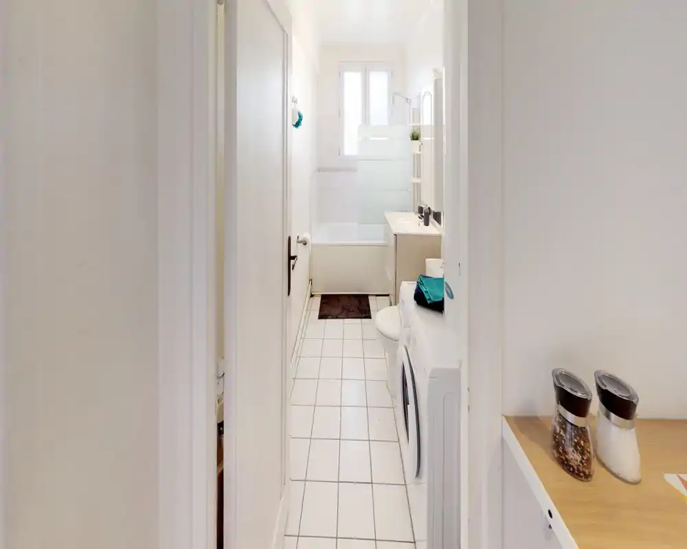 Bagno stretto e luminoso con vasca, mobile lavabo e specchio a parete; vicino all’ingresso si vede una lavatrice. Piastrelle bianche e luce naturale dalla finestra rendono l’ambiente arioso.