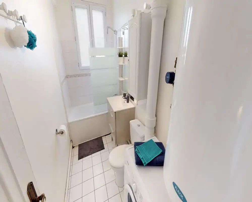 Bagno compatto e pulito con vasca/doccia, mobile lavabo con specchio, WC e lavatrice. Piastrelle chiare e piccoli dettagli decorativi rendono lo spazio funzionale per gli ospiti.