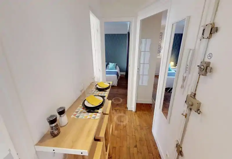 Spazio comune luminoso e stretto con un tavolo a parete pieghevole apparecchiato per due e vista su una camera decorata; pavimento in legno e arredo moderno ideale per un appartamento compatto.