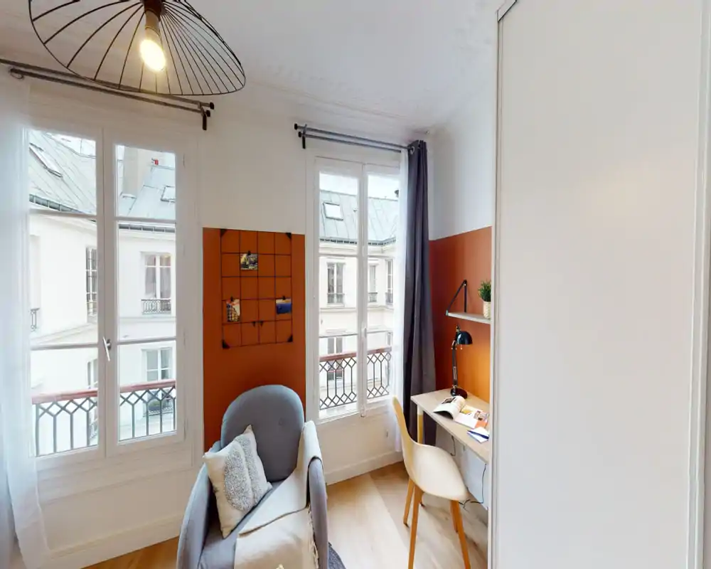 Bright, well-staged living corner with a comfortable armchair, desk and large windows letting in natural light — ideal for relaxing or working.