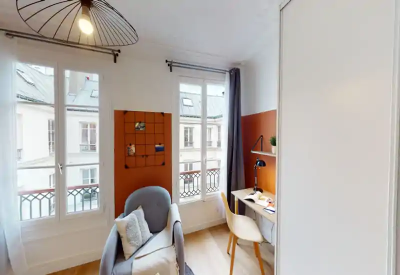 Bright, well-staged living corner with a comfortable armchair, desk and large windows letting in natural light — ideal for relaxing or working.