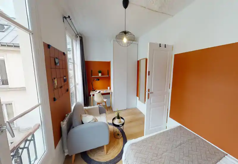 Bright, stylish bedroom with a comfortable bed partially visible, a cozy armchair, a small desk and wardrobe — ideal for a single renter. Large windows provide excellent natural light and the warm accent wall adds character.