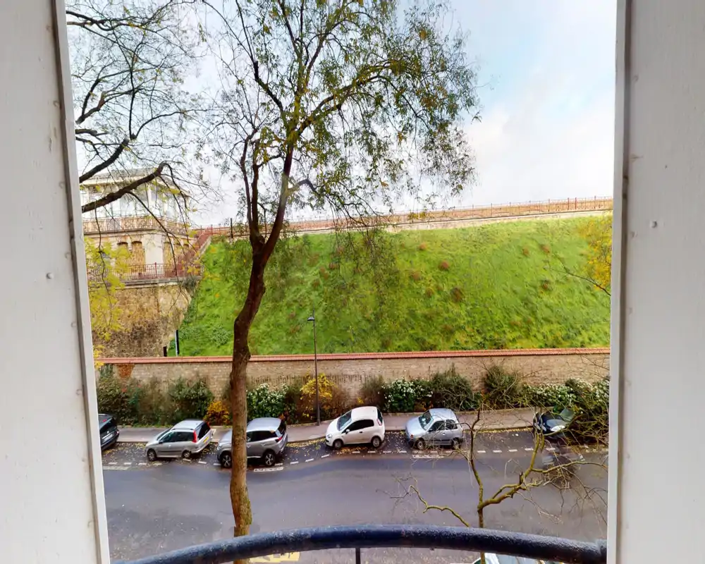 Balcony view overlooking a tree-lined street and a grassy embankment; ideal for morning coffee with street parking visible below.