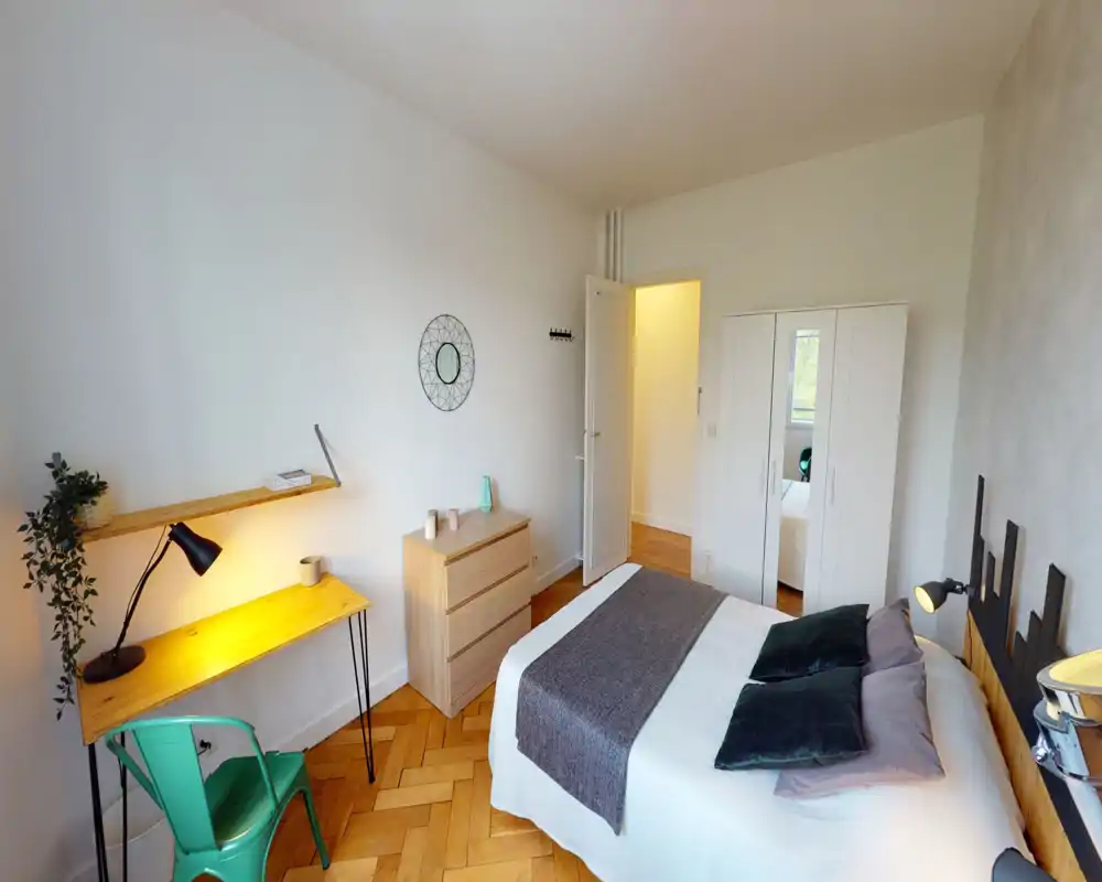 Bright, modern bedroom with a comfortable double bed, soft textiles and a wooden headboard. A small desk and chair by the window add a practical workspace while white walls and parquet flooring create a warm, airy atmosphere.