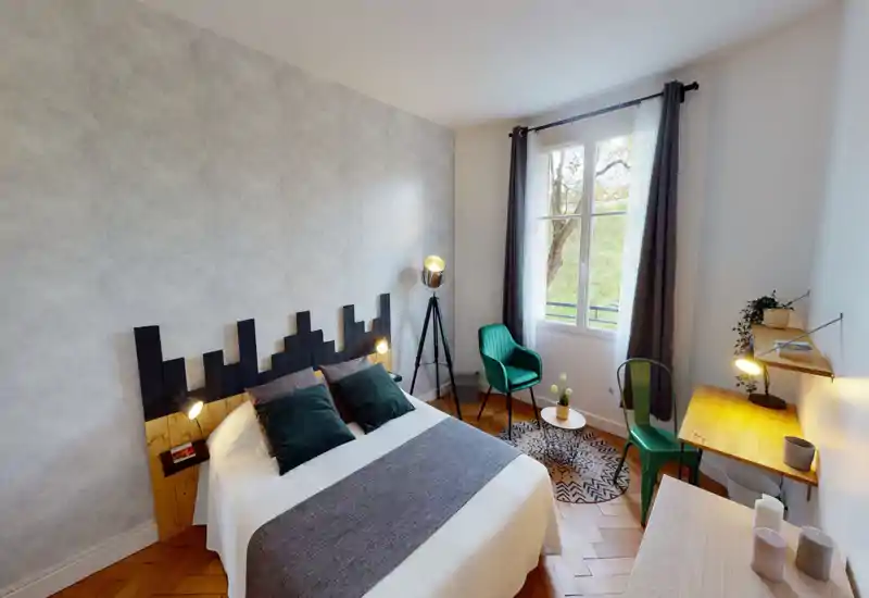 Bright, stylish bedroom with a double bed, decorative headboard and coordinated green accents. A window fills the room with natural light and a small seating area with a rug and chair creates a cozy corner.