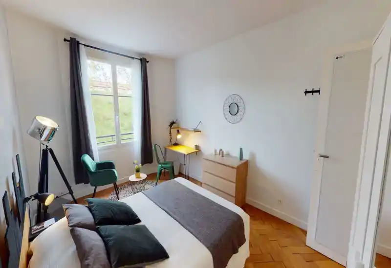 Bright, stylish bedroom with a neatly made double bed, soft textiles and a cozy reading corner by the window. A wooden dresser and a small desk with a lamp complete the clean, modern layout.