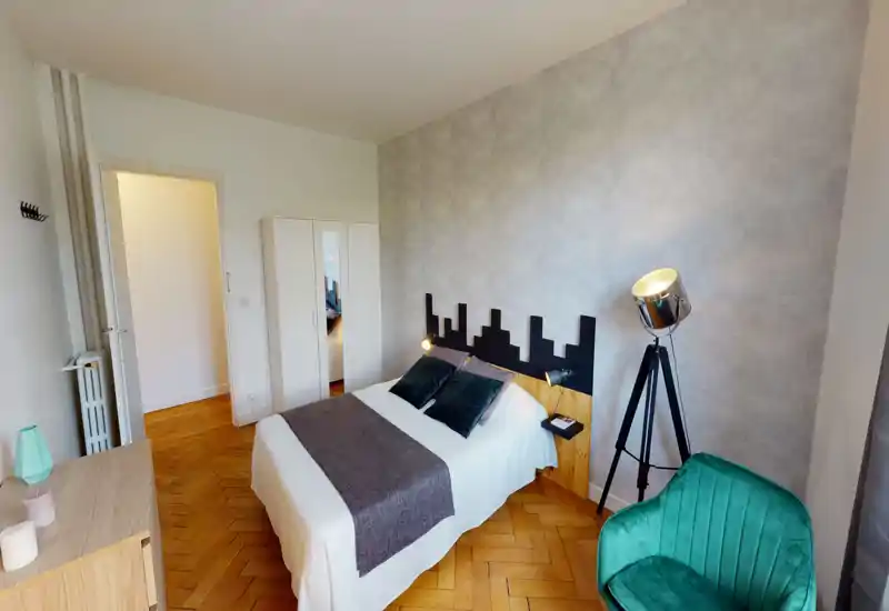 Bright, well-styled bedroom with a double bed, decorative headboard and soft textiles. The room features a wardrobe, tripod floor lamp and a comfortable green armchair, with warm parquet flooring and neutral textured walls.