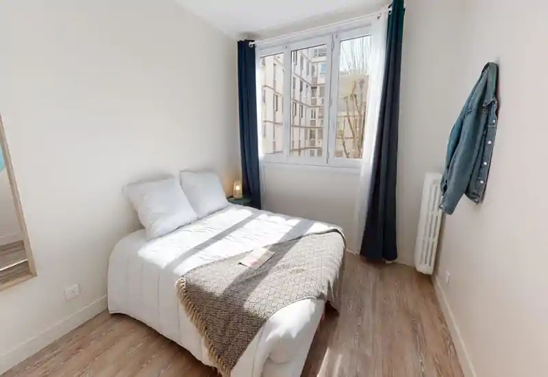 Chambre lumineuse et ensoleillée avec un lit double bien fait, linge blanc et un plaid texturé. Grande fenêtre avec rideaux foncés apportant une belle lumière naturelle et vue sur la ville.