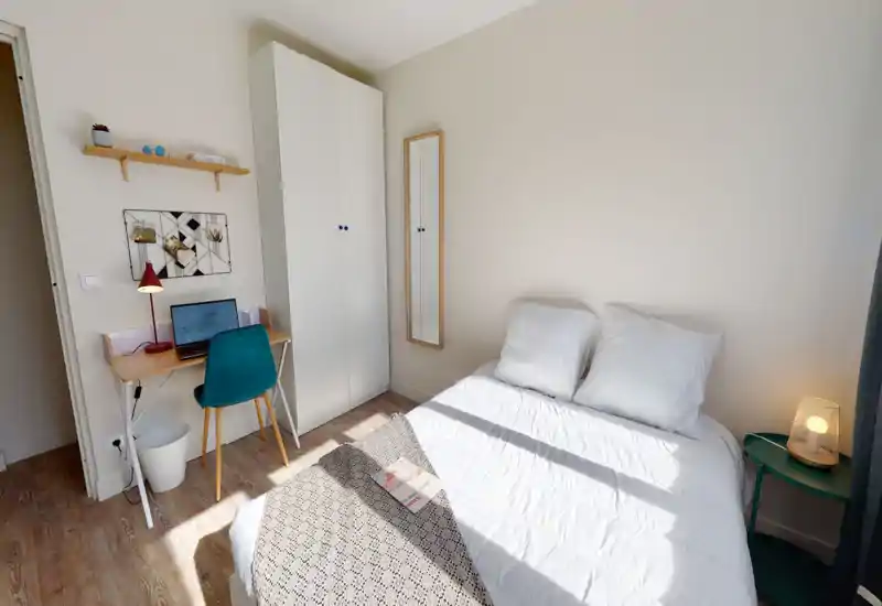 Chambre lumineuse et soigneusement mise en scène avec un lit double confortable, une lampe de chevet et un petit espace bureau. La lumière naturelle illumine le sol en bois et la décoration minimaliste.