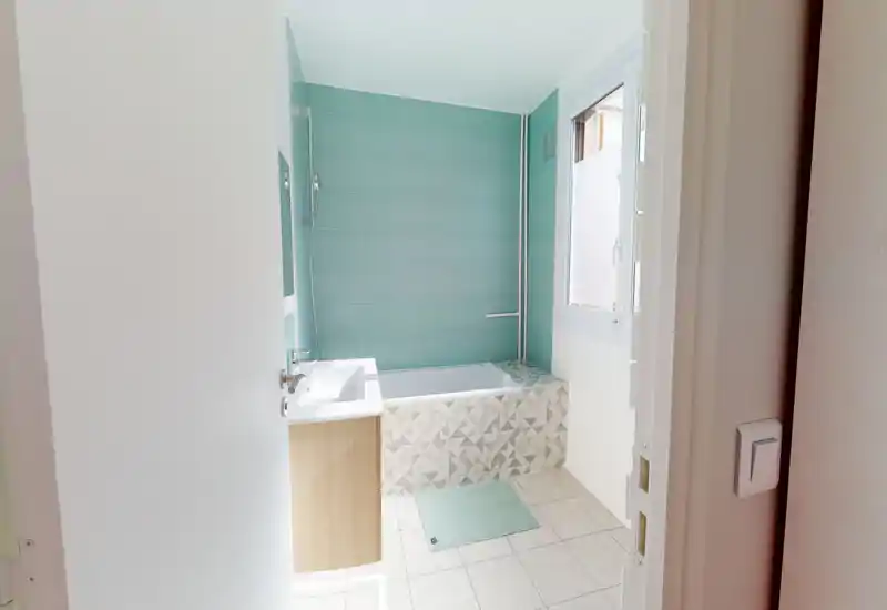 Salle de bain moderne et propre avec baignoire, mur carrelé turquoise et lavabo avec meuble en bois. Espace lumineux avec motifs géométriques sur la façade de la baignoire et une fenêtre apportant la lumière naturelle.