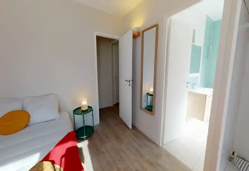Chambre compacte et lumineuse avec un lit bien fait, une lampe de chevet chaleureuse et un sol en bois ; une porte donne sur une salle de bains lumineuse aux carreaux turquoise.