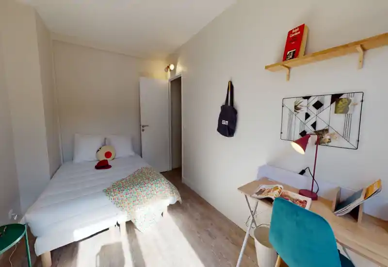 Chambre simple lumineuse et épurée, lit bien fait avec coussin décoratif et petit bureau près de la fenêtre. Sol en bois clair et murs neutres apportent une atmosphère calme et accueillante.