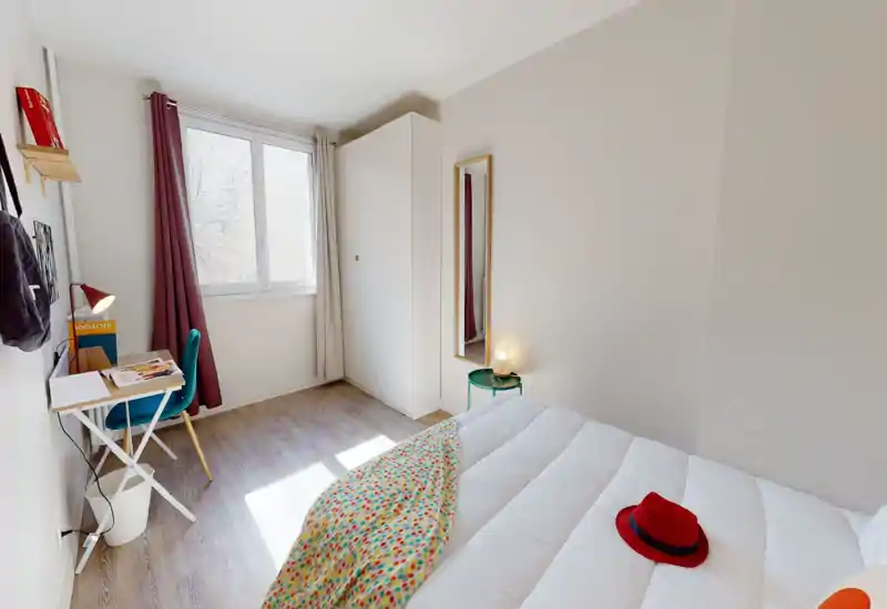 Chambre moderne et lumineuse avec lit blanc, jeté coloré et chapeau rouge sur la couette ; grande fenêtre, bureau et armoire pour un séjour fonctionnel.