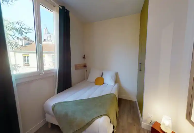 Camera da letto compatta e luminosa con un letto matrimoniale ben fatto sotto la finestra, tessili morbidi e una lampada da comodino che creano un’atmosfera accogliente.