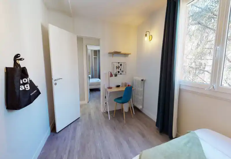 Angolo di camera luminoso e moderno con una scrivania piccola e il letto parzialmente visibile vicino alla finestra; pavimento in legno chiaro, sedia turchese e applique creano un ambiente accogliente e funzionale.