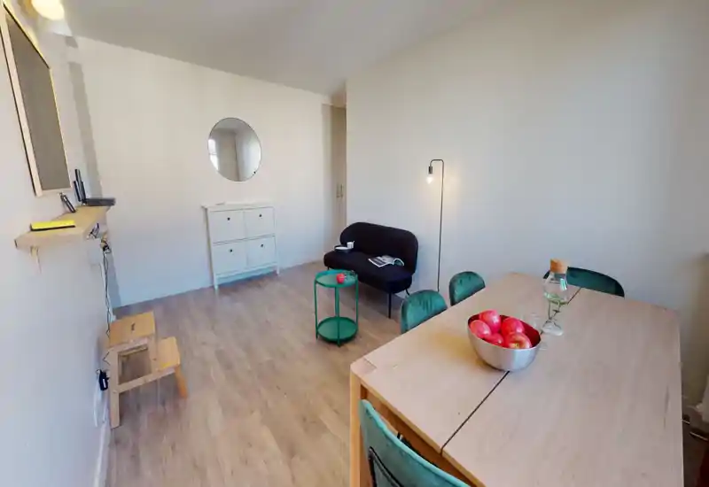 Séjour lumineux et minimaliste avec un petit canapé sombre, une table d'appoint verte et une table à manger en bois avec chaises vertes ; sol net et miroir rond au mur pour une ambiance moderne et aérée.