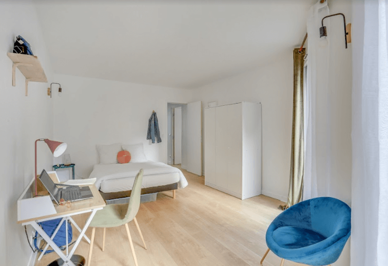 Chambre lumineuse et minimaliste avec lit double, lampe de chevet, armoire et un petit bureau — idéale pour un séjour confortable au style moderne et aéré.