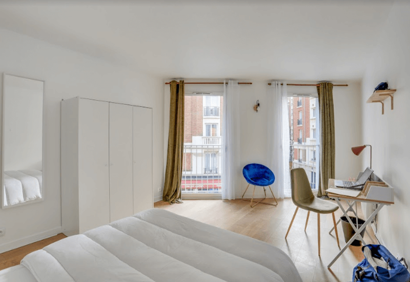 Chambre lumineuse et moderne avec grand lit, armoire, bureau et deux grandes fenêtres apportant beaucoup de lumière naturelle. Décor minimaliste, sol en bois chaleureux et fauteuil d'appoint confortable.