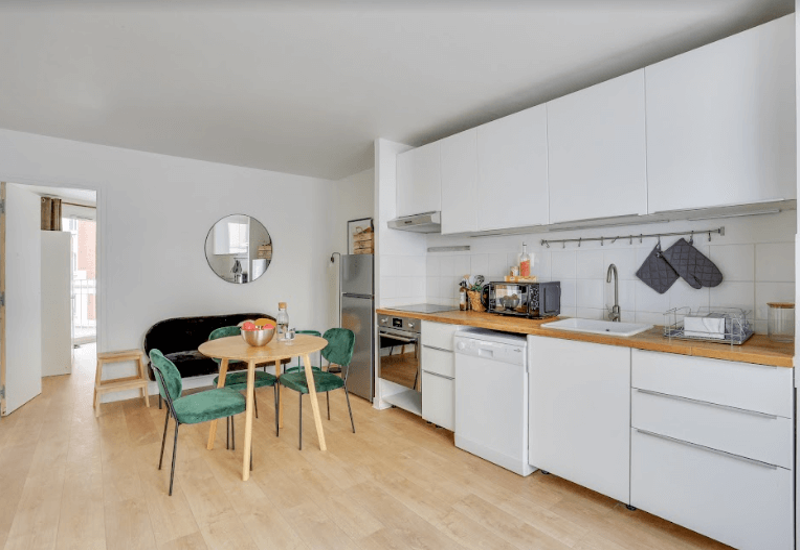 Cuisine ouverte moderne et lumineuse avec placards blancs, plan de travail en bois et petite table à manger avec chaises vertes — propre, bien équipée et idéale pour un appartement compact.