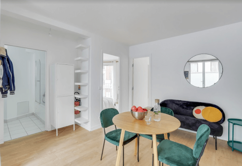 Espace de vie moderne et lumineux avec une table à manger ronde en bois et quatre chaises en velours, un petit canapé aux coussins colorés et un miroir mural qui reflète la lumière naturelle.